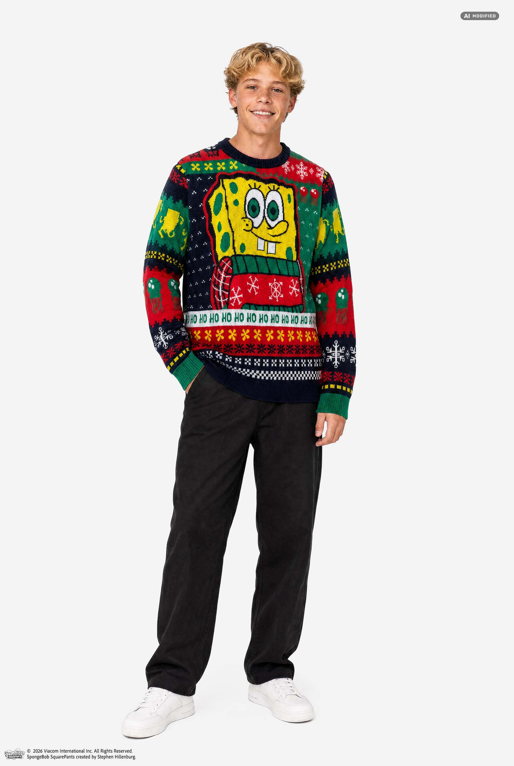 A young man wearing a SpongeBob-themed Christmas sweater, standing against a plain light gray studio background. The sweater features a large SpongeBob graphic on the chest and festive multicolor holiday patterns including snowflakes, bells, and 'HO HO HO' text.