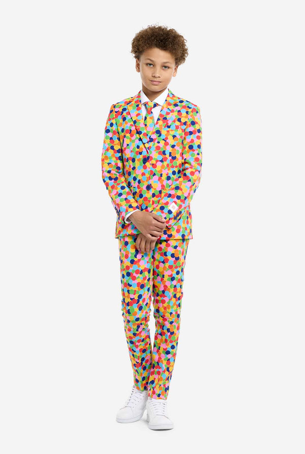 In this image, a kid is wearing a colorful, polka-dotted suit. The suit has a bright pattern with circles in various colors including orange, yellow, pink, green, blue, and red, arranged in a confetti-like style. Hands in front of suit.
