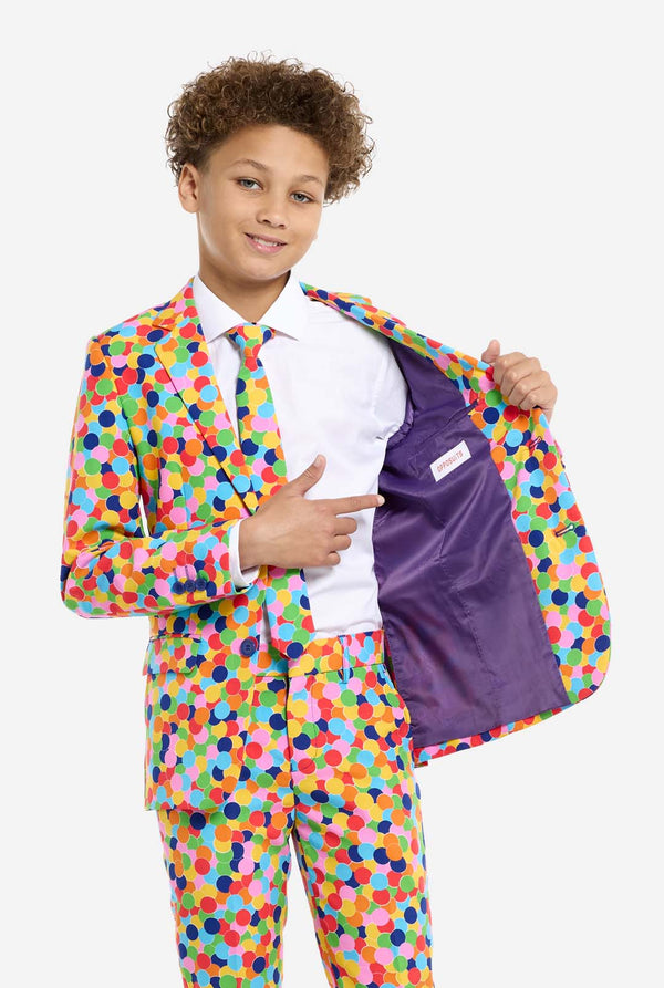 In this image, a kid is wearing a colorful, polka-dotted suit. The suit has a bright pattern with circles in various colors including orange, yellow, pink, green, blue, and red, arranged in a confetti-like style. Holding one side of the suit open showing purple inside