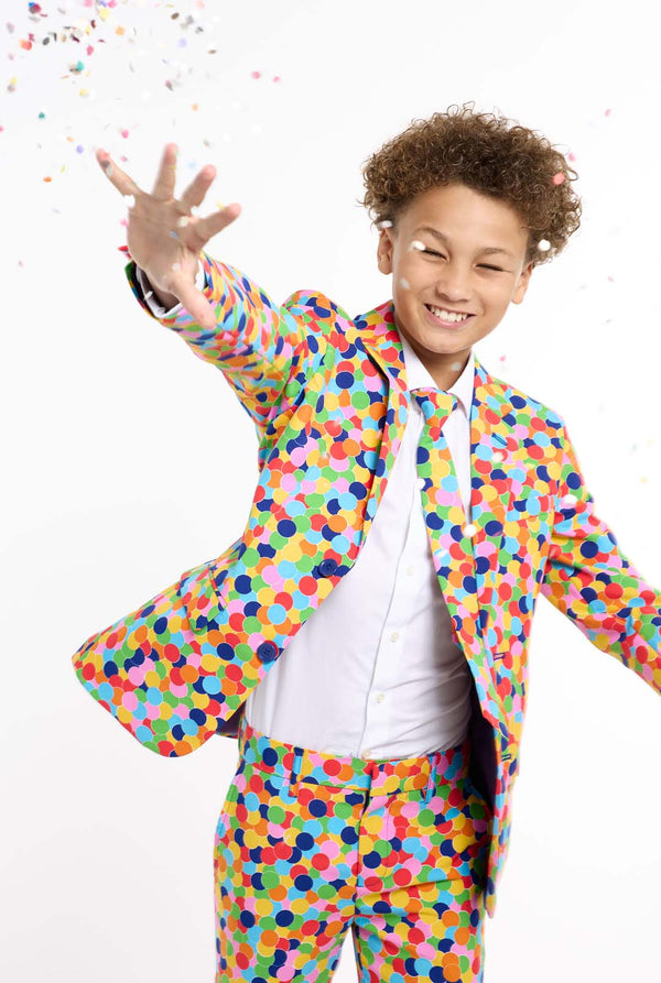 In this image, a kid is wearing a colorful, polka-dotted suit. The suit has a bright pattern with circles in various colors including orange, yellow, pink, green, blue, and red, arranged in a confetti-like style. Trowing confetti with one hand open.