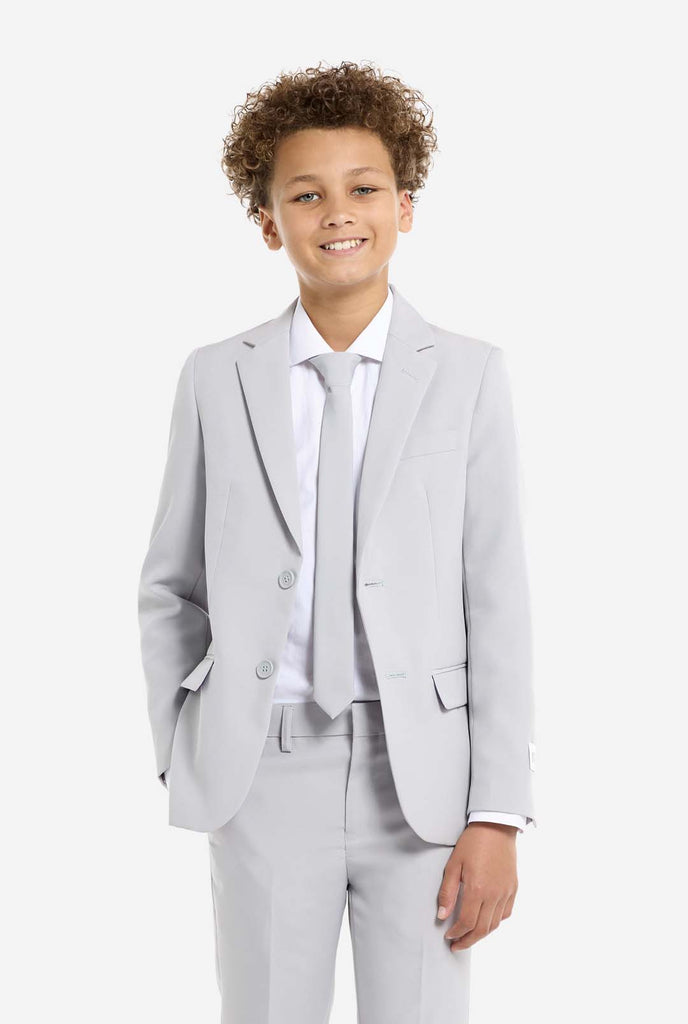 In this image, a kid is wearing a formal grey suit. The suit consists of a blazer, pants and tie. Kid has one hand in pocket.