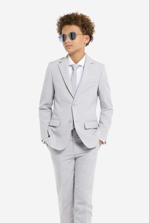 In this image, a kid is wearing a formal grey suit and sunglasses. The suit consists of a blazer, pants and tie. Kid has hands in pocket.