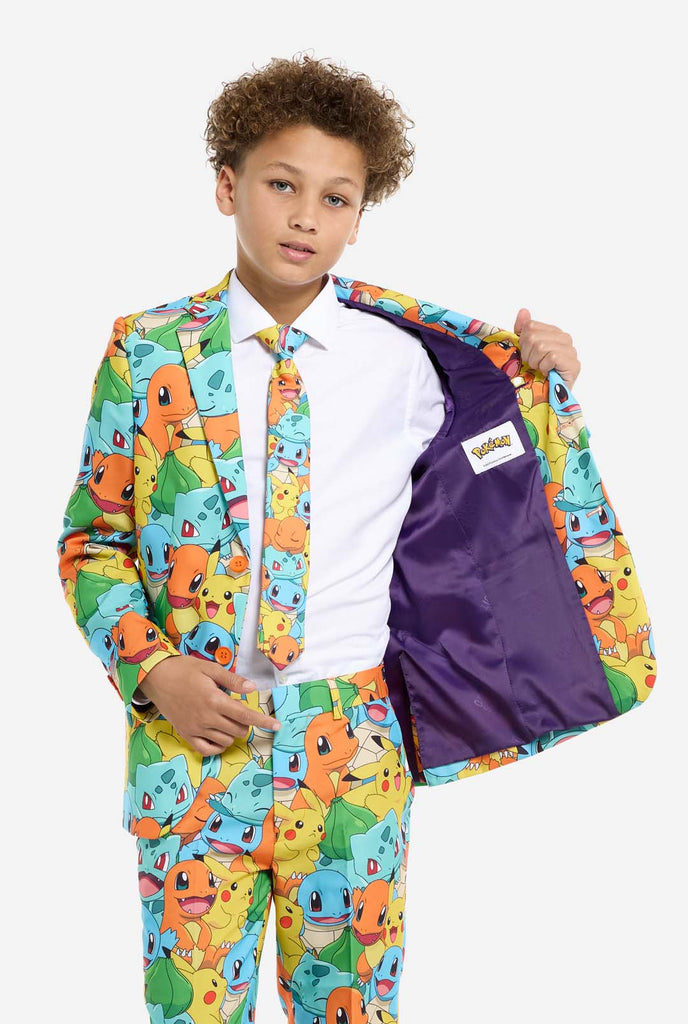 Teen wearing formal multi color suit with all-over Pokemon print. The suit contains the characters Pikachu, Bulbasaur, Charmander and Squirtle. Holding one side of the jacket open.