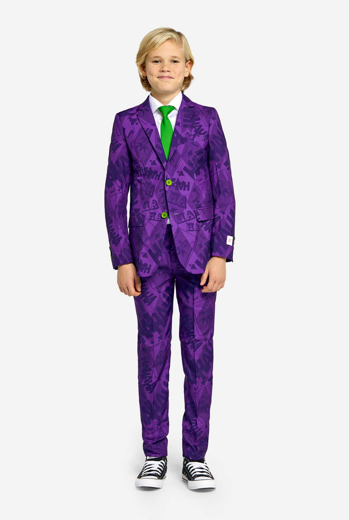 Teen wearing purple teen boys suit with a The Joker theme and green buttons. Further he is wearing a green tie, a white shirt and black and white sneakers. Arms aside his body.