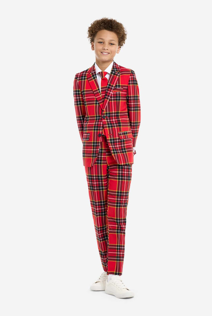 A young boy is wearing a stylish red plaid suit, known as The Lumberjack by OppoSuits. This teen boys' suit features a bold red, black, and yellow tartan pattern, perfect for holiday parties, formal events, or casual occasions that call for a playful yet classy look. The suit includes a matching jacket, trousers, and tie, all designed with a slim fit cut for a modern, youthful appearance. The boy pairs the suit with a crisp white shirt, enhancing the colors of the plaid pattern. Holding hands behind back.