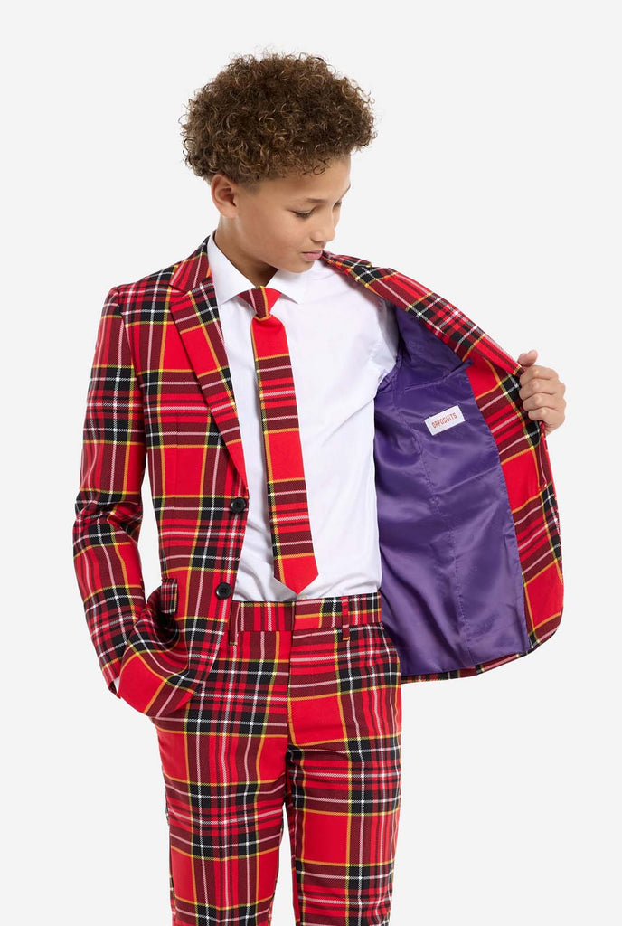 A young boy is wearing a stylish red plaid suit, known as The Lumberjack by OppoSuits. This teen boys' suit features a bold red, black, and yellow tartan pattern, perfect for holiday parties, formal events, or casual occasions that call for a playful yet classy look. The suit includes a matching jacket, trousers, and tie, all designed with a slim fit cut for a modern, youthful appearance. The boy pairs the suit with a crisp white shirt and holding one side of the jacket open.