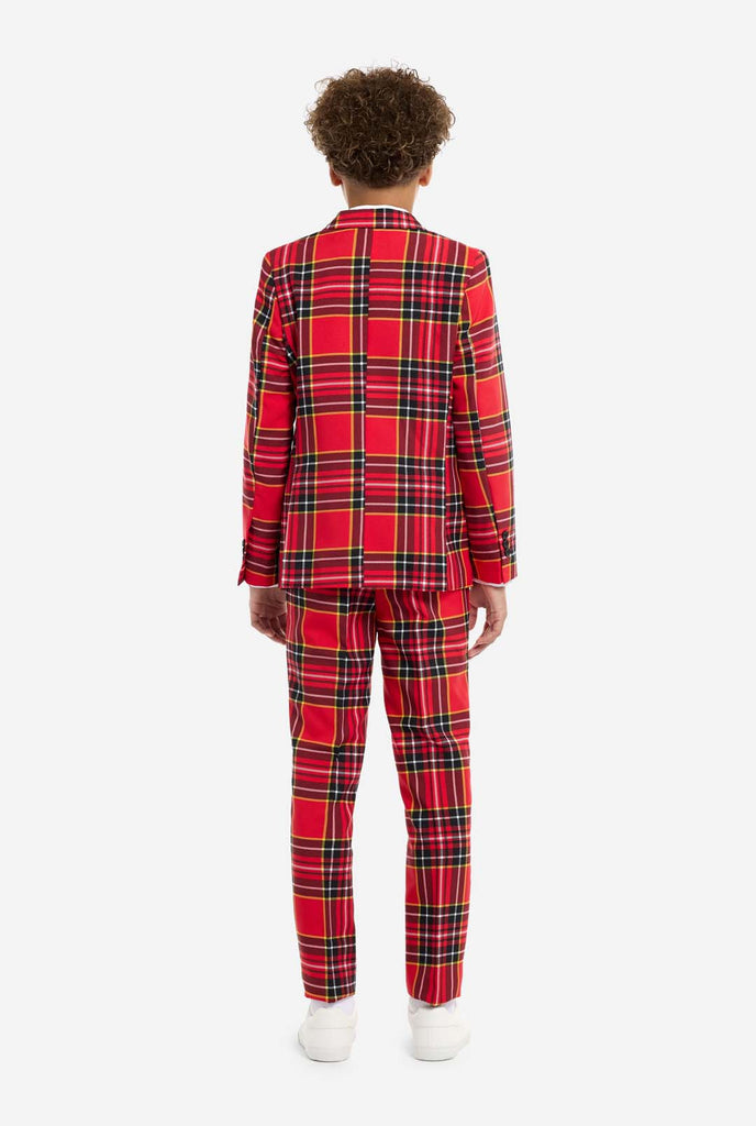 A young boy is wearing a stylish red plaid suit, known as The Lumberjack by OppoSuits. This teen boys' suit features a bold red, black, and yellow tartan pattern, perfect for holiday parties, formal events, or casual occasions that call for a playful yet classy look. The suit includes a matching jacket, trousers, and tie, all designed with a slim fit cut for a modern, youthful appearance. The boy pairs the suit with a crisp white shirt, enhancing the vibrant colors of the plaid pattern. View from the back.