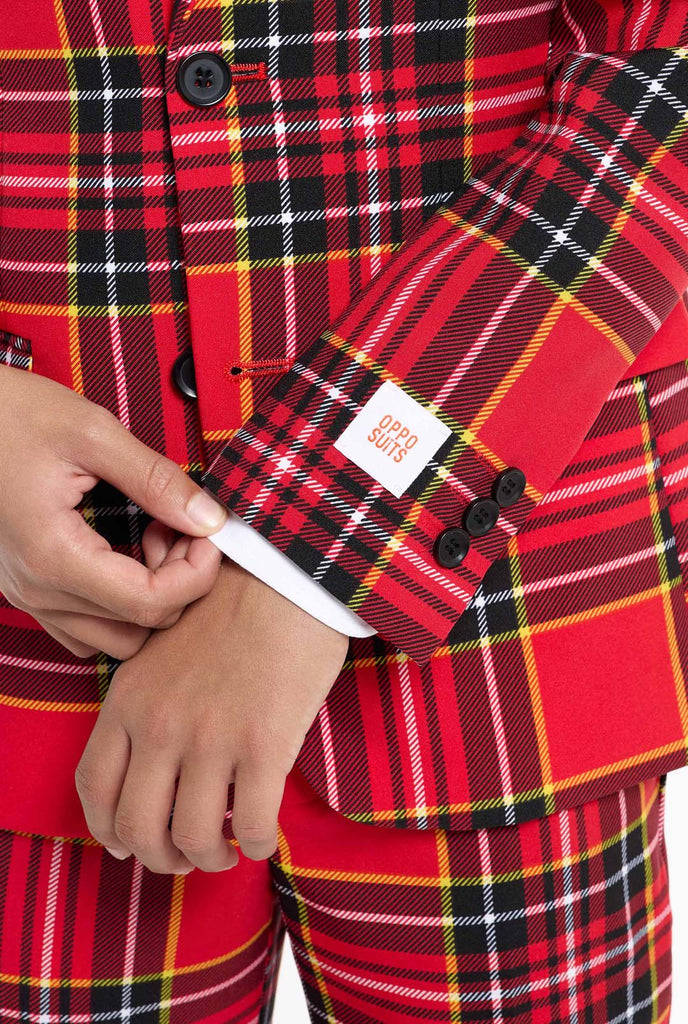 A young boy is wearing a stylish red plaid suit, known as The Lumberjack by OppoSuits. This teen boys' suit features a bold red, black, and yellow tartan pattern, perfect for holiday parties, formal events, or casual occasions that call for a playful yet classy look. The suit includes a matching jacket, trousers, and tie, all designed with a slim fit cut for a modern, youthful appearance. The boy pairs the suit with a crisp white shirt, enhancing the vibrant colors of the plaid pattern. Closeup sleeve.