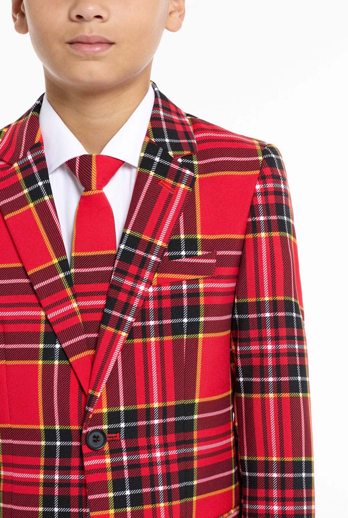 A young boy is wearing a stylish red plaid suit, known as The Lumberjack by OppoSuits. This teen boys' suit features a bold red, black, and yellow tartan pattern, perfect for holiday parties, formal events, or casual occasions that call for a playful yet classy look. The suit includes a matching jacket, trousers, and tie, all designed with a slim fit cut for a modern, youthful appearance. The boy pairs the suit with a crisp white shirt, enhancing the vibrant colors of the plaid pattern. Chest close up.