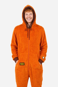 Male model wearing an orange South Park Kenny adult onesie with brown hood trim and sleeve patch. With hands in the pocket.