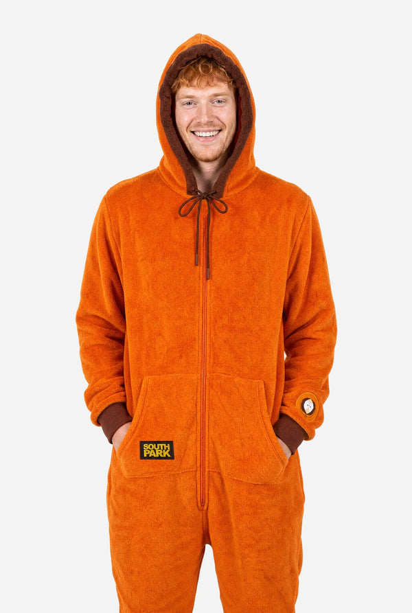 Male model wearing an orange South Park Kenny adult onesie with brown hood trim and sleeve patch. With hands in the pocket.