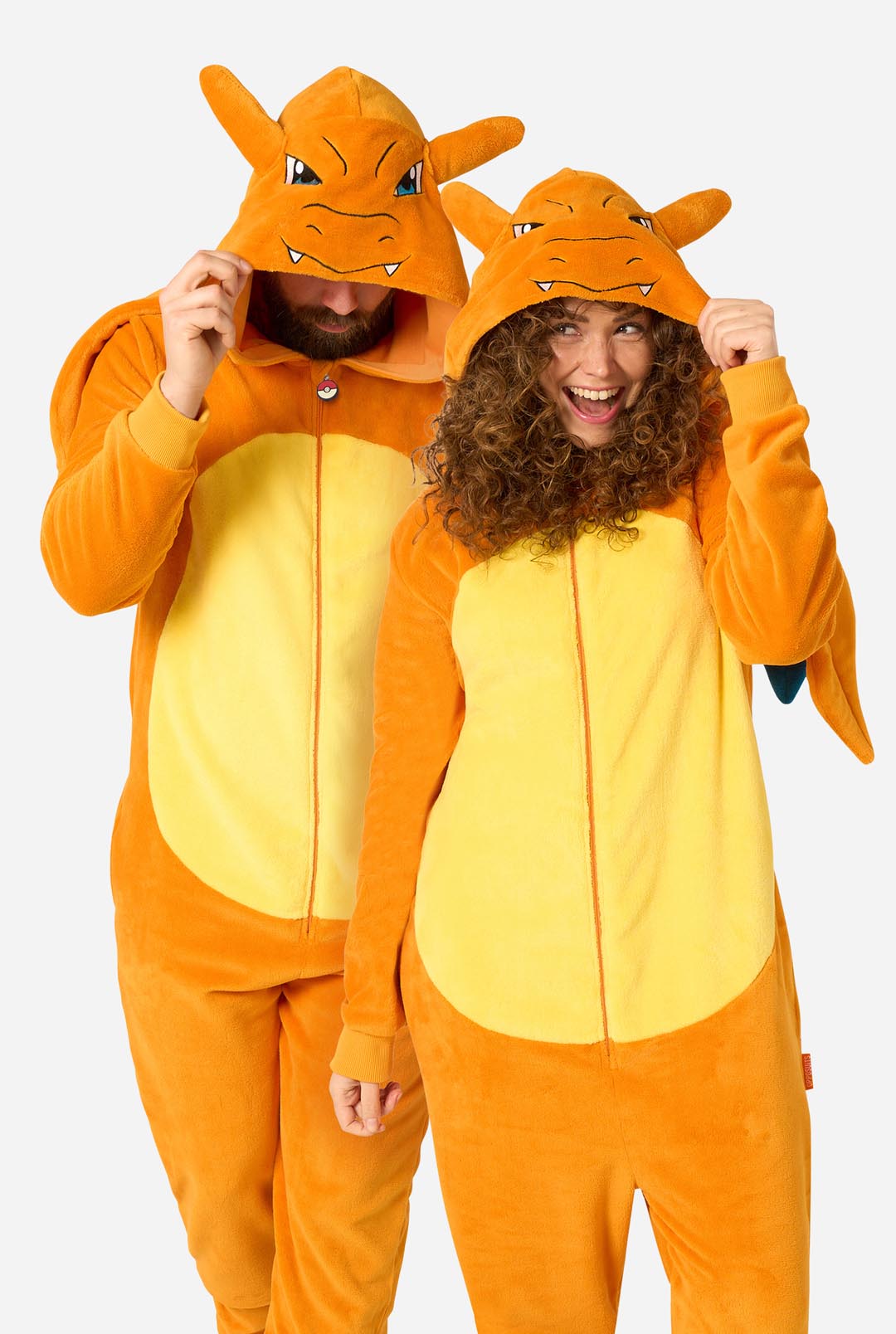 Two people wearing matching orange and yellow Charizard-themed hooded onesies against a plain light gray background.