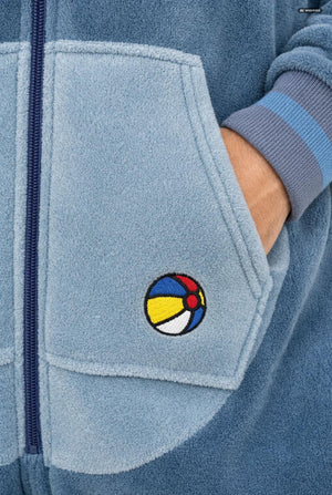 Close-up of a light blue fleece zip-up jacket with a front pocket featuring a colorful embroidered beach ball patch and a striped ribbed cuff.