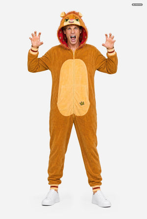 A man modeling a brown plush lion costume onesie with a hood, featuring a large beige belly patch on the front, orange cuffs and collar trim, and white sneakers, set against a plain light gray background.