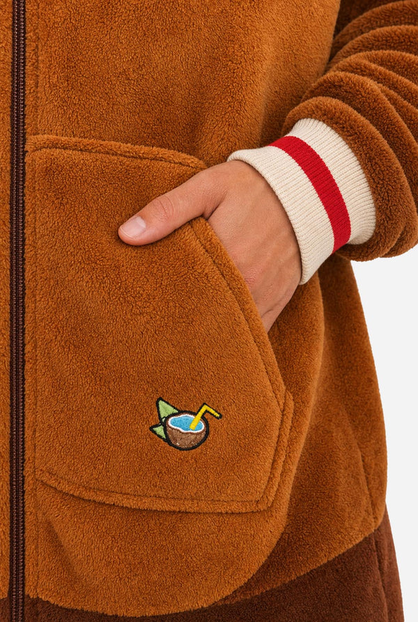 A man wearing a brown plush animal onesie with a hood shaped like a monkey face, featuring pink ears, a yellow inner hood lining, and a large tan belly patch on the front, photographed against a white studio background. Close up of the pocket.