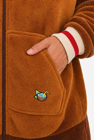 Close-up of a person wearing a warm brown fleece zip-up jacket, showing a hand in a front pocket with a small embroidered cocktail graphic and a cream ribbed cuff with a red stripe.