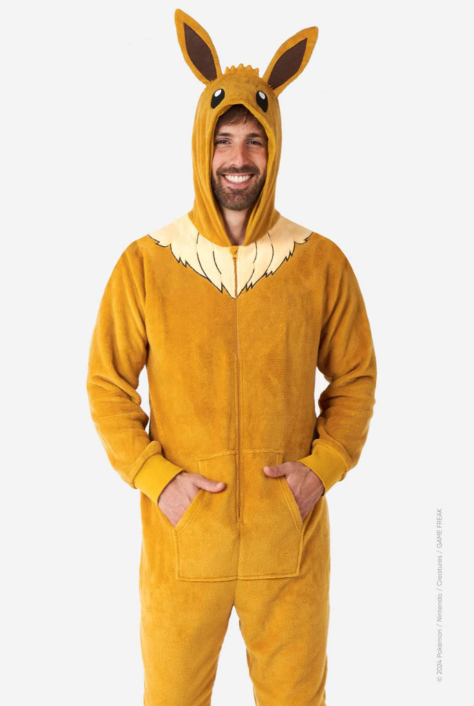 A man is wearing an adorable Eevee onesie, modeled after the beloved Pokémon character. This adult unisex onesie is designed in a warm brown color, resembling Eevee’s fur, and features a fluffy beige collar mimicking Eevee’s mane. The hood includes Eevee's signature large brown ears, big black eyes. The onesie has a front zipper closure and convenient pockets at the front, offering both comfort and ease of wear. Perfect for Pokémon fans.