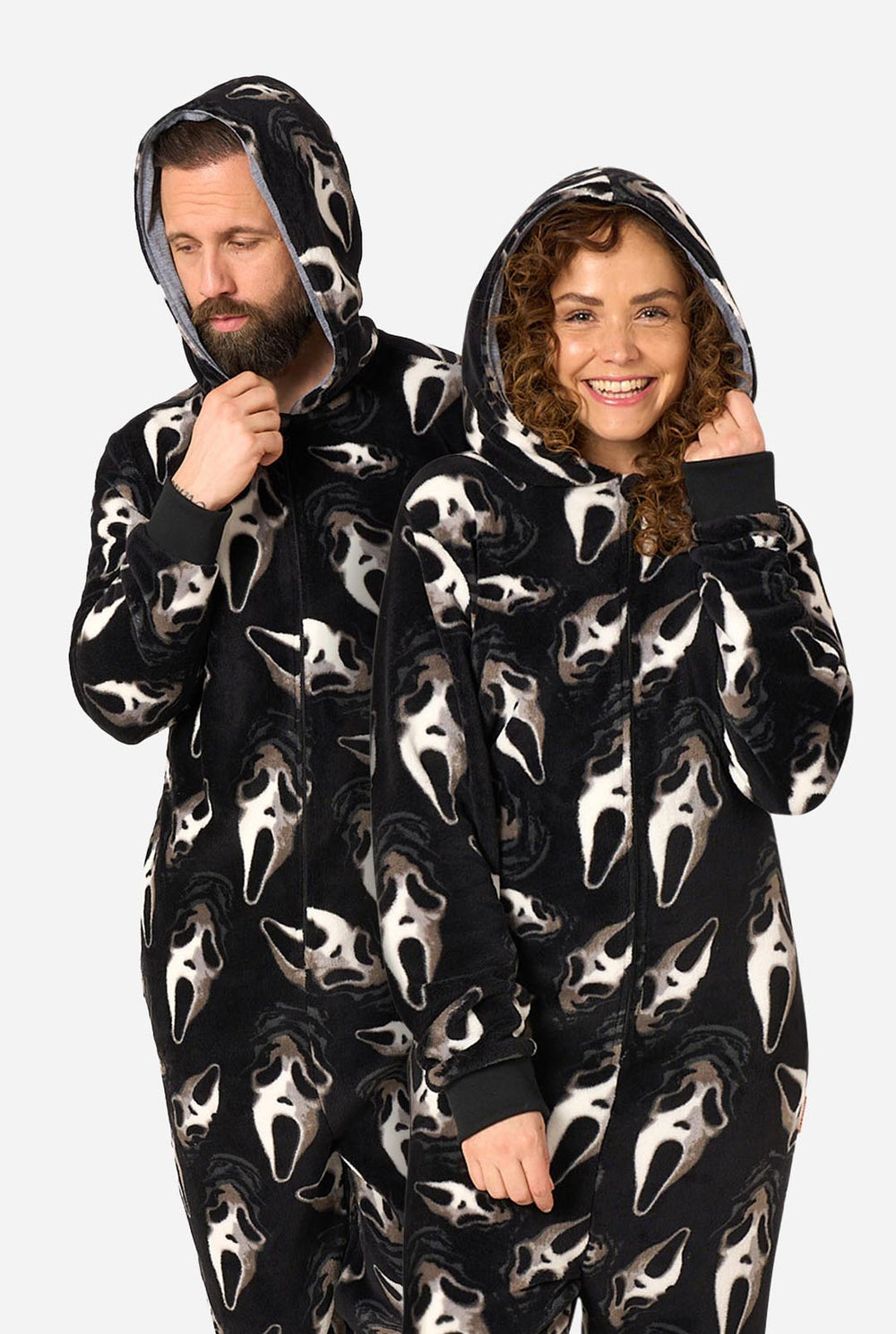 A man and a woman wearing matching black hooded onesies with an all-over Ghostface mask print, posed against a light gray studio background.