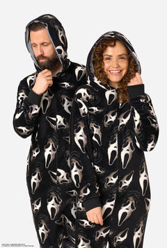 A man and a woman modeling matching black hooded onesie pajamas with an all-over white Ghost Face print, standing against a light gray studio background.