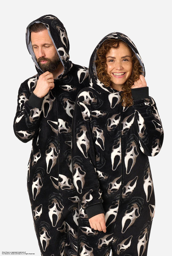 A man and a woman modeling matching black hooded onesie pajamas with an all-over white Ghost Face print, standing against a light gray studio background.