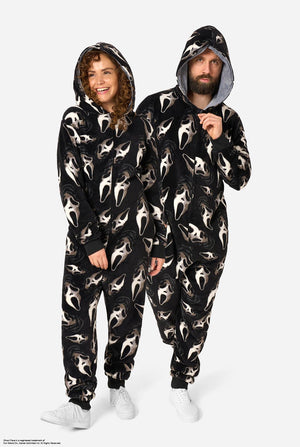 A man and a woman modeling matching black hooded onesie pajamas with an all-over white Ghost Face print, standing against a light gray studio background. Full-body image.