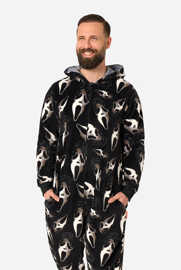 A smiling man with a beard wearing a black hooded one-piece pajama or kigurumi with an all-over Ghostface face print, posed against a light gray studio background.
