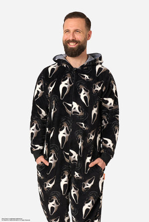 A man modeling black hooded onesie pajamas with an all-over white Ghost Face print, standing against a light gray studio background.