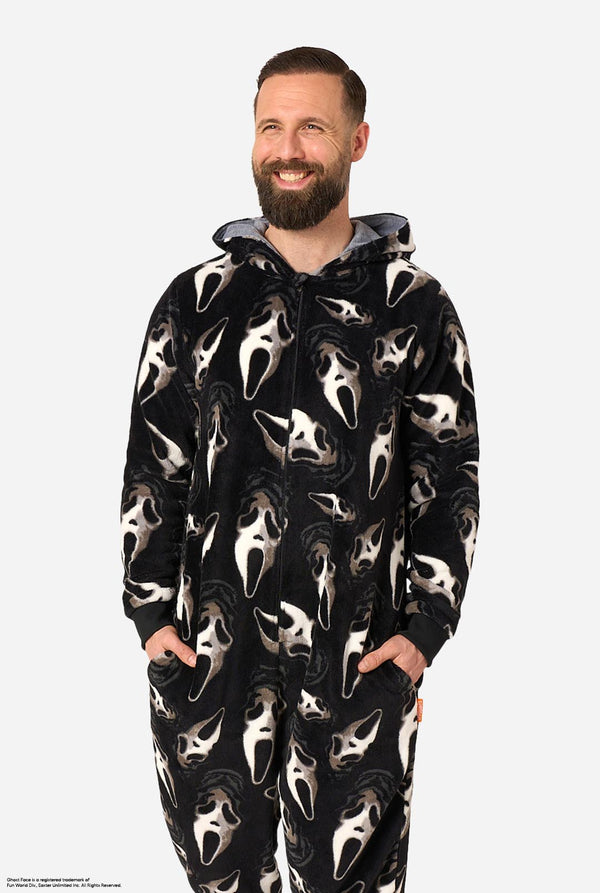 A man modeling black hooded onesie pajamas with an all-over white Ghost Face print, standing against a light gray studio background.