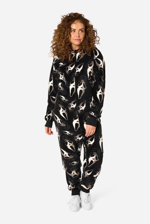 A model with curly brown hair wearing a black hooded one-piece pajama suit with a white screaming face pattern, paired with white sneakers, against a light gray background.