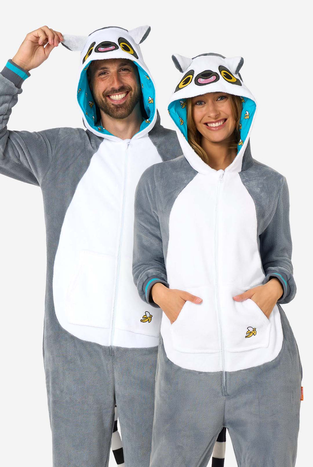 A man and women are wearing a grey and white lemur adult onesie featuring playful hood details and cozy, soft material, perfect for lounging or costume events. Including black and white tail. The hood are on their heads.