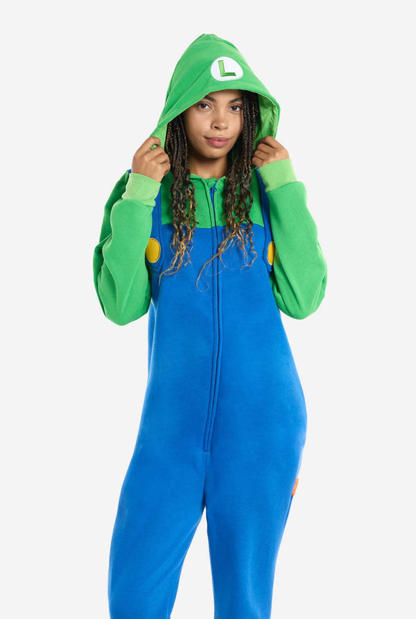 A woman is dressed in a Luigi-themed onesie. The outfit features a bright green hoodie with an "L" symbol on the hood and blue overalls with large yellow buttons on the straps. She is holding both sides of the hood, as if pulling it over her head, and looking directly at the camera with a neutral expression. The overall design mimics the look of the popular video game character Luigi, with contrasting green and blue colors. The background is plain, putting focus on the costume and the woman's playful pose.
