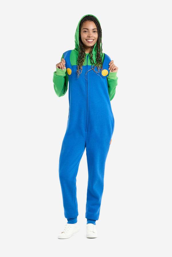 A woman is dressed in a Luigi-themed onesie. The outfit features a bright green hoodie with an "L" symbol on the hood and blue overalls with large yellow buttons on the straps. She is holding the straps and looking directly at the camera with a neutral expression. The overall design mimics the look of the popular video game character Luigi, with contrasting green and blue colors. The background is plain, putting focus on the costume and the woman's playful pose.