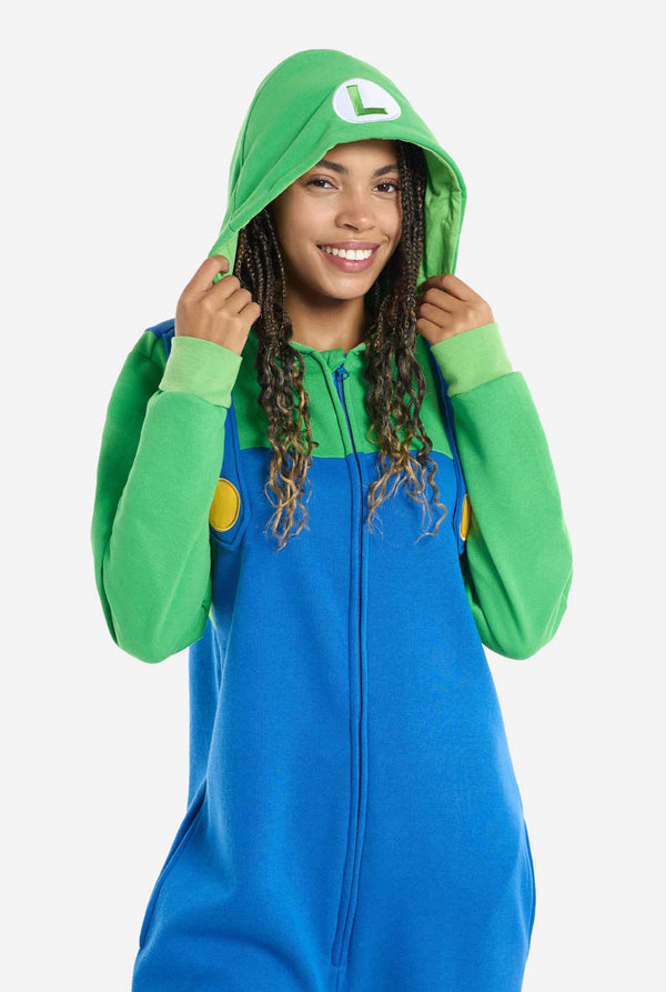A woman is dressed in a Luigi-themed onesie. The outfit looks as a bright green hoodie with an "L" symbol on the hood and blue overalls with large yellow buttons on the straps. She is holding both sides of the hood, as if pulling it over her head, and looking directly at the camera. The overall design mimics the look of the popular video game character Luigi, with contrasting green and blue colors. The background is plain, putting focus on the costume and the woman's pose. Zoomed image.