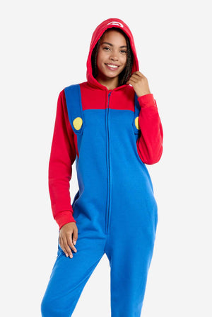 A woman is dressed in a Mario-themed onesie. The outfit features a bright red hoodie with an "M" symbol on the hood and blue overalls with large yellow buttons on the straps. She is holding one side of the hood, as if pulling it over her head, and looking directly at the camera with a neutral expression. The overall design mimics the look of the popular video game character Mario, with contrasting red and blue colors. The background is plain, putting focus on the costume. 