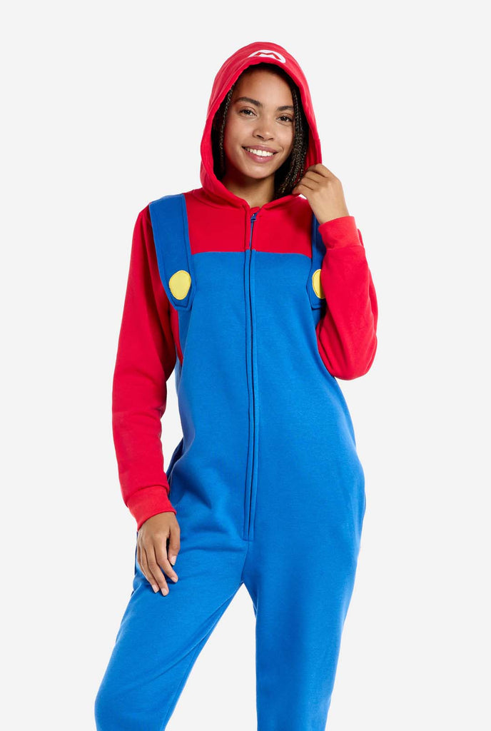 A woman is dressed in a Mario-themed onesie. The outfit features a bright red hoodie with an "M" symbol on the hood and blue overalls with large yellow buttons on the straps. She is holding one side of the hood, as if pulling it over her head, and looking directly at the camera with a neutral expression. The overall design mimics the look of the popular video game character Mario, with contrasting red and blue colors. The background is plain, putting focus on the costume. 