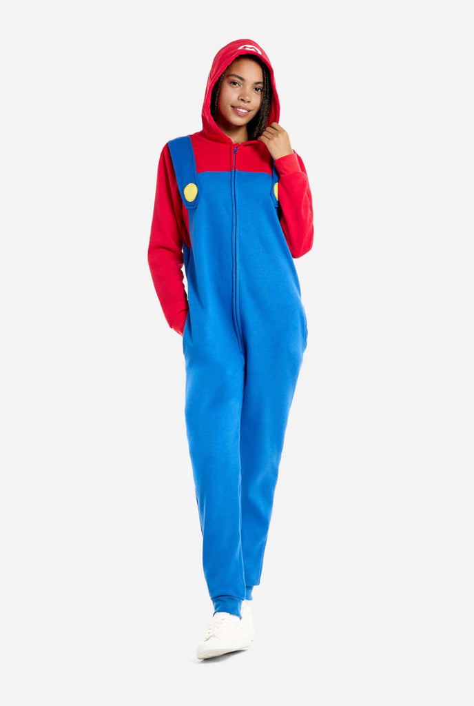 A woman is dressed in a Mario-themed onesie. The outfit features a bright red hoodie with an "M" symbol on the hood and blue overalls with large yellow buttons on the straps. She is holding one side of the hood, as if pulling it over her head, and looking directly at the camera with a neutral expression. The overall design mimics the look of the popular video game character Mario, with contrasting red and blue colors. The background is plain, putting focus on the costume and the woman's playful pose.