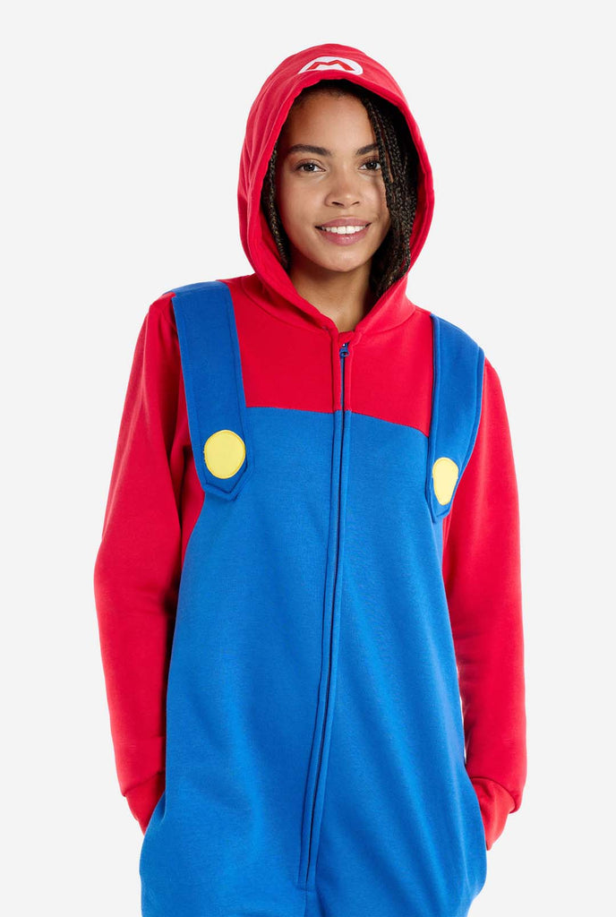 A woman is dressed in a Mario-themed onesie. The outfit looks as a bright red hoodie with an "M" symbol on the hood and blue overalls with large yellow buttons on the straps. Hands in the pockets and looking directly at the camera. The overall design mimics the look of the popular video game character Mario, with contrasting red and blue colors. The background is plain, putting focus on the costume and the woman's pose. Zoomed image.