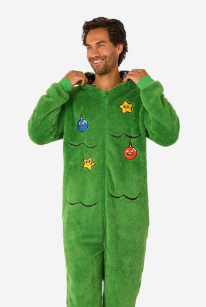 A smiling man wearing a green Christmas onesie designed to look like a Christmas tree, decorated with cartoon ornaments and stars.