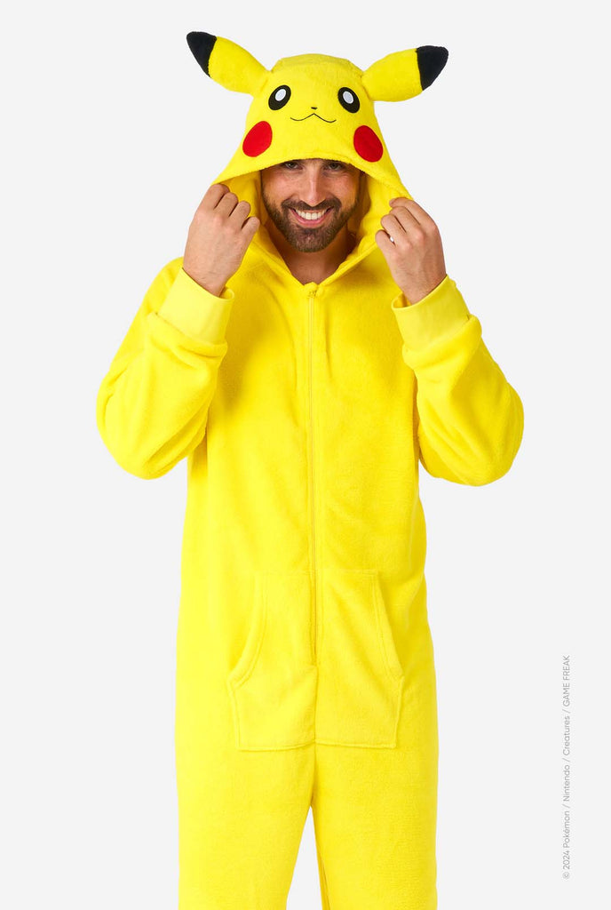 A man is wearing a vibrant Pikachu onesie, designed to resemble the iconic Pokémon character. This unisex adult onesie is bright yellow with Pikachu’s recognizable face on the hood, featuring large black-tipped ears, black eyes, red cheeks, and a cute smile. The onesie has a convenient front zipper closure and two front pockets for functionality and comfort. The soft, cozy material makes it perfect for lounging, cosplay events, or Pokémon-themed gatherings. Ideal for Pokémon fans.