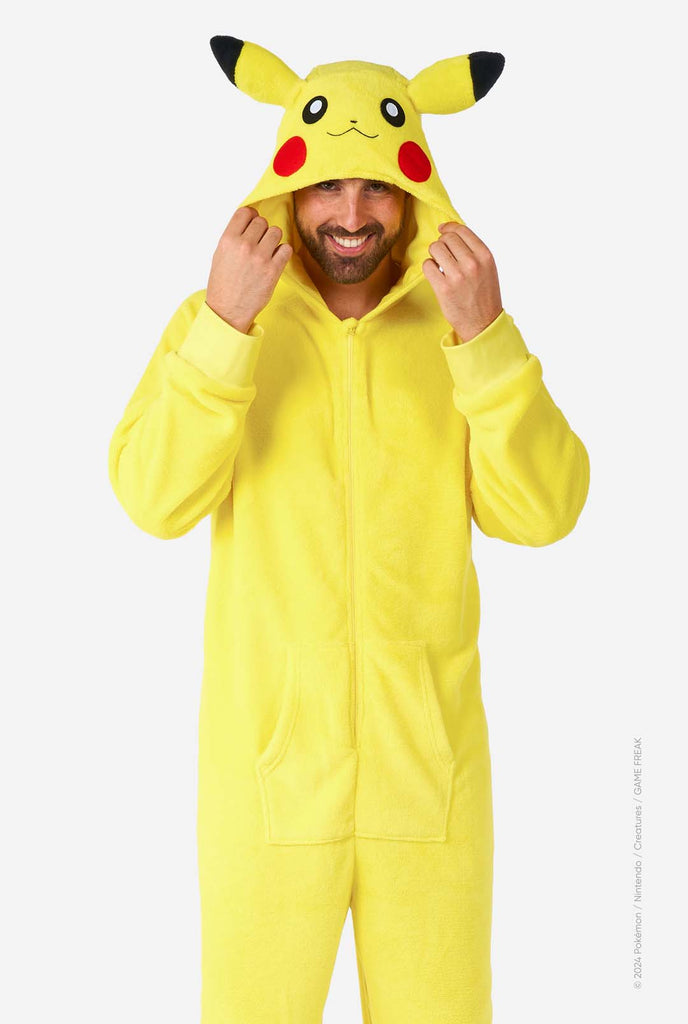 A man is wearing a vibrant Pikachu onesie, designed to resemble the iconic Pokémon character. This unisex adult onesie is bright yellow with Pikachu’s recognizable face on the hood, featuring large black-tipped ears, black eyes, red cheeks, and a cute smile. The onesie has a convenient front zipper closure and two front pockets for functionality and comfort. The soft, cozy material makes it perfect for lounging, cosplay events, or Pokémon-themed gatherings. Ideal for Pokémon fans.