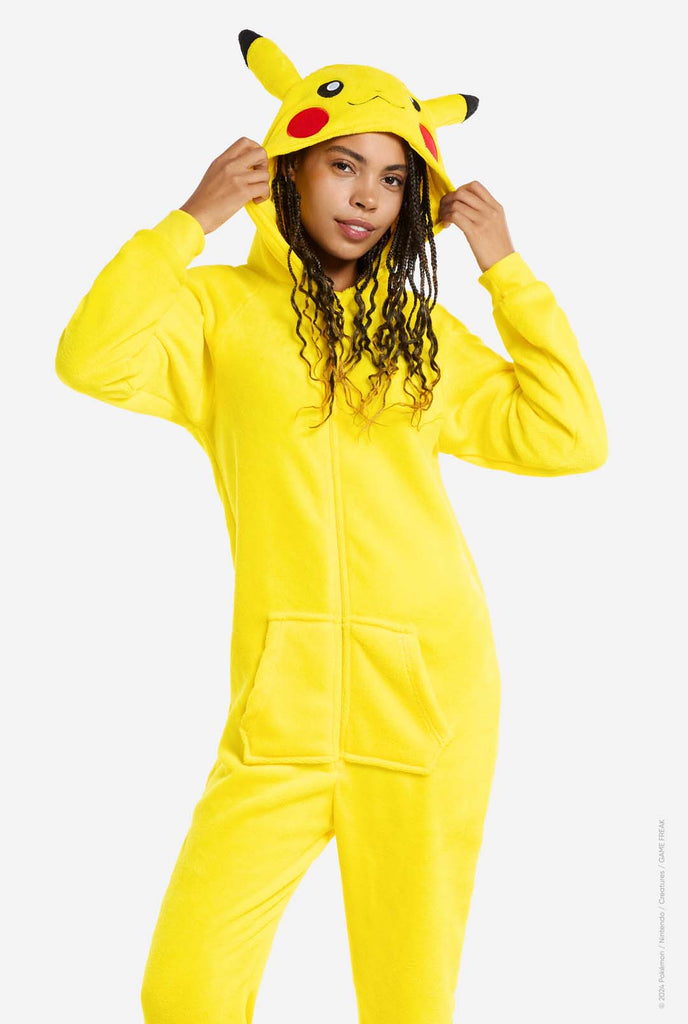 A woman is wearing a vibrant Pikachu onesie, designed to resemble the iconic Pokémon character. This unisex adult onesie is bright yellow with Pikachu’s recognizable face on the hood, featuring large black-tipped ears, black eyes, red cheeks, and a cute smile. The onesie has a convenient front zipper closure and two front pockets for functionality and comfort. The soft, cozy material makes it perfect for lounging, cosplay events, or Pokémon-themed gatherings. Ideal for Pokémon fans.