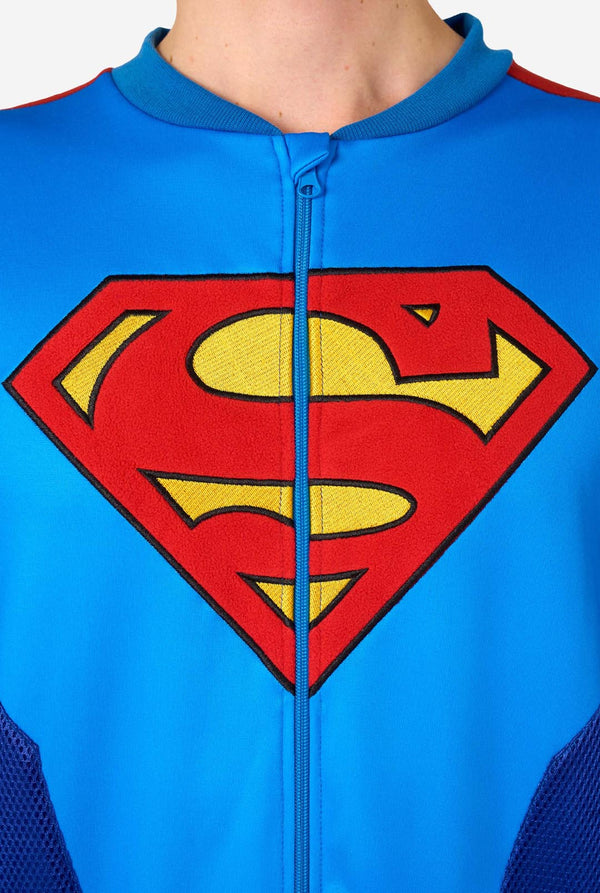 Image of a man wearing a blue and red Superman onesie with a red cape on a light background. Chest logo close up.
