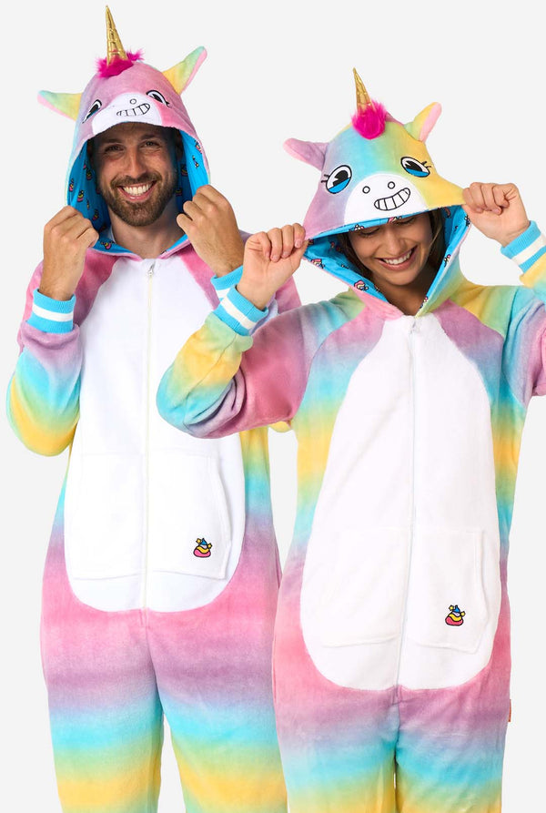 A man and women are wearing a colorful adult onesie designed as a rainbow unicorn. The onesie features a vibrant gradient of pastel colors, a golden horn, and playful unicorn details, perfect for cozy lounging or costume parties. The men and women both have their hood on.