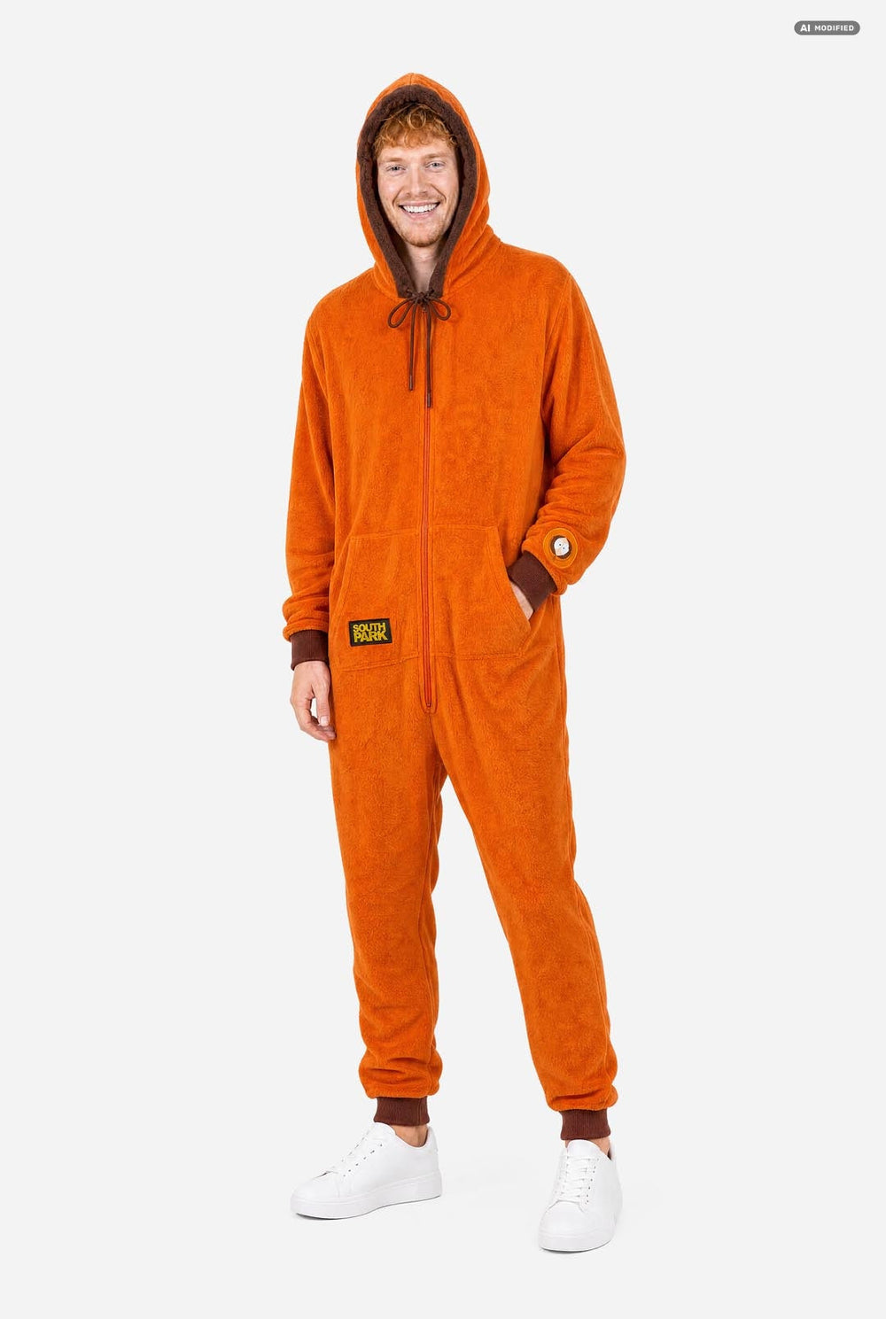 A smiling man wearing an orange South Park Kenny onesie with brown drawstrings and cuffs, standing against a light gray background. A small patch on the left leg reads 'South Park'.  
