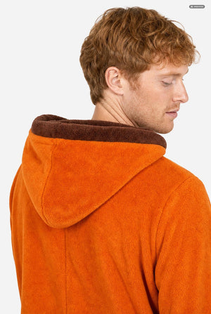 A smiling man wearing an orange South Park Kenny onesie with brown drawstrings and cuffs, standing against a light gray background. A small patch on the left leg reads 'South Park'.  Hood close up.
