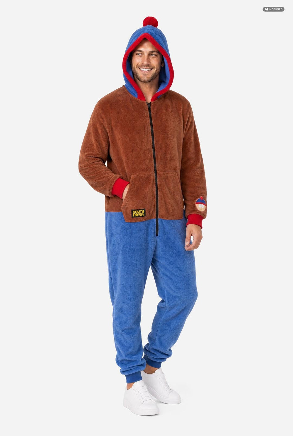 A man wearing a brown and blue South Park Stan onesie with blue and red hood lining, and a South Park patch on the sleeve, photographed against a light gray studio background.