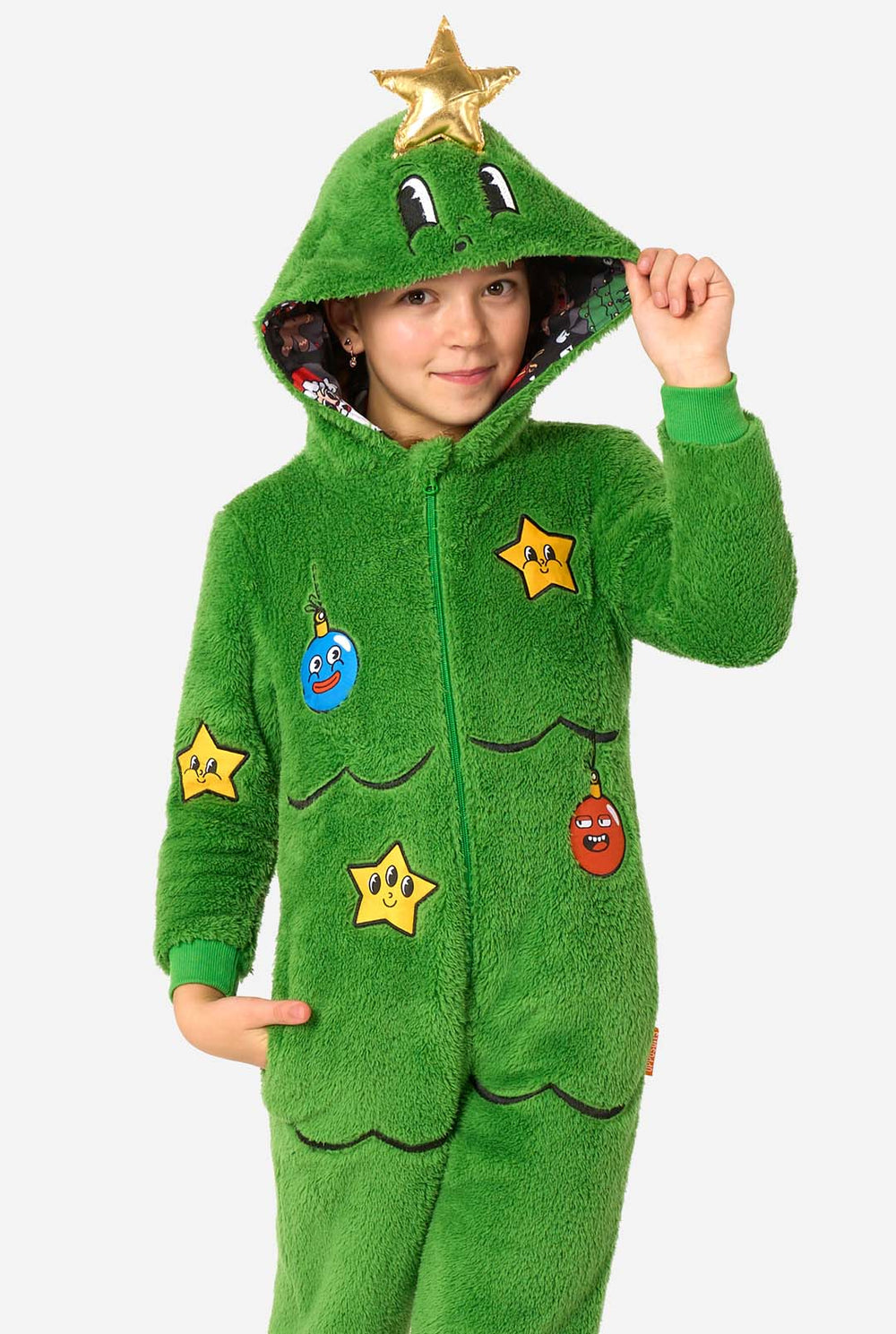 Child wearing a green Christmas tree–shaped onesie with a hood featuring cartoon eyes and a gold star on top, holding the hood.
