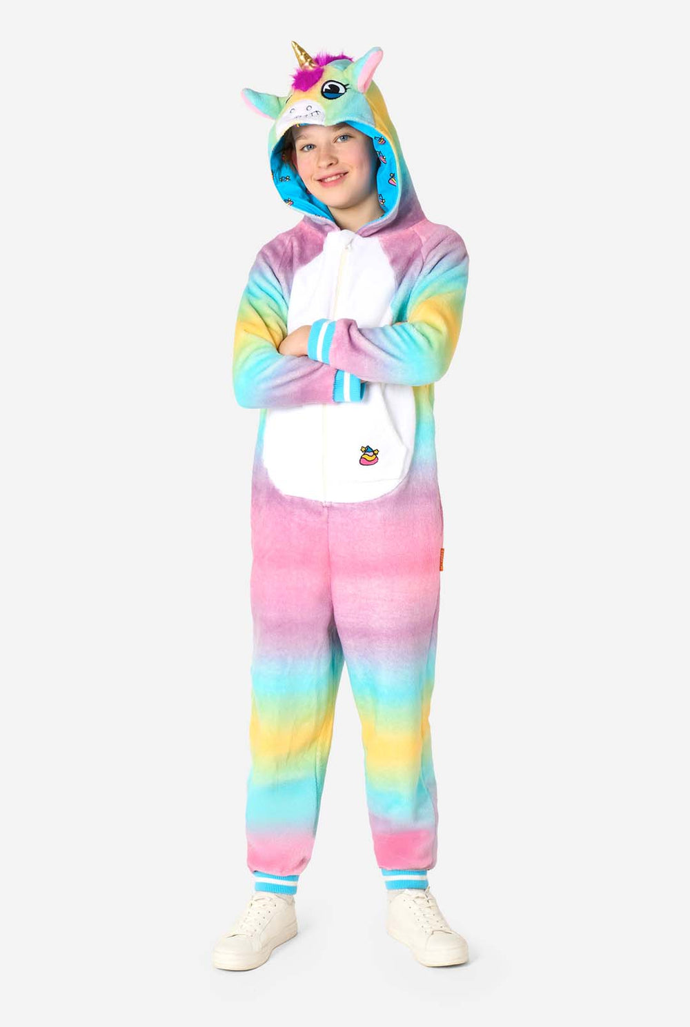 A smiling kid is wearing a white, pink, blue and yellow kids onesie designed as a unicorn. The Unicorn onesie features a hood resembling a unicorn head and other playfull details