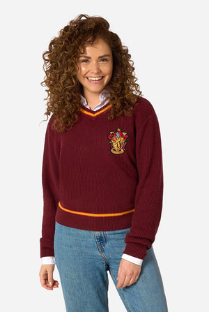 A woman with curly brown hair wearing a maroon ribbed knit Harry Potter Gryffindor sweater with gold and yellow trim, featuring the crest of Gryffindor on the left chest, paired with blue jeans against a light gray background.