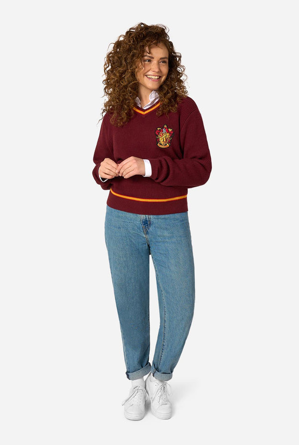 A woman modeling a maroon long-sleeve crewneck Harry Potter Gryffindor sweater with yellow and white striped trim and a Gryffindor crest on the chest, paired with blue jeans and white sneakers, against a light gray background.